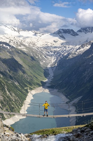 Mountaineers on a suspension bridge over a mountain stream Alelebach, picturesque mountain landscape near the Olpererhuette, view of turquoise-blue lake Schlegeisspeicher, glaciated rocky mountain peaks Grosser Moeseler, Hoher Weisszint and Hochfeilermit glacier Schlegeiskees, Berliner Hoehenweg, Zillertal Alps, Tyrol, Austria, Europe