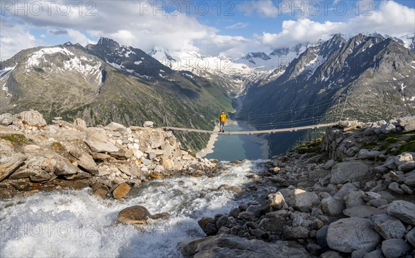 Mountaineers on a suspension bridge over a mountain stream Alelebach, picturesque mountain landscape near the Olpererhuette, view of turquoise-blue lake Schlegeisspeicher, glaciated rocky mountain peaks Grosser Moeseler, Hoher Weisszint and Hochfeilermit glacier Schlegeiskees, Berliner Hoehenweg, Zillertal Alps, Tyrol, Austria, Europe