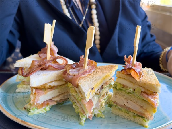 Woman Sitting Around a Table in Restaurant and Eating Club Sandwich with Bacon in a Sunny Day in Switzerland