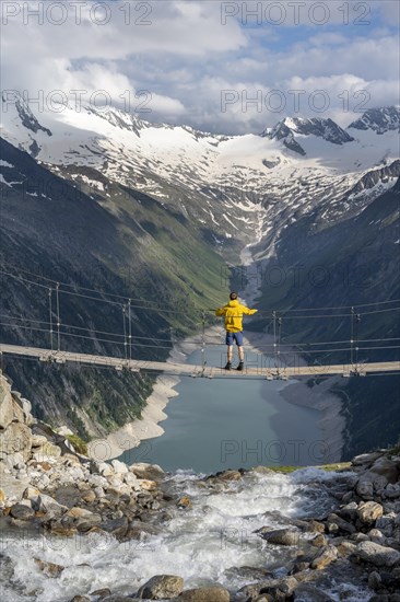 Mountaineers on a suspension bridge over a mountain stream Alelebach, picturesque mountain landscape near the Olpererhuette, view of turquoise-blue lake Schlegeisspeicher, glaciated rocky mountain peaks Grosser Moeseler, Hoher Weisszint and Hochfeiler with glacier Schlegeiskees, Berliner Hoehenweg, Zillertal Alps, Tyrol, Austria, Europe