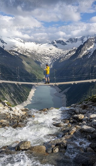 Mountaineers on a suspension bridge over a mountain stream Alelebach, picturesque mountain landscape near the Olpererhuette, view of turquoise-blue lake Schlegeisspeicher, glaciated rocky mountain peaks Hoher Weisszint and Hochfeiler with glacier Schlegeiskees, Berliner Hoehenweg, Zillertal Alps, Tyrol, Austria, Europe