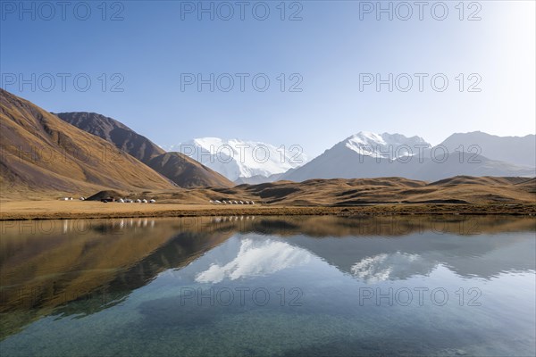 White glaciated and snow-covered mountain peak Pik Lenin at sunset, mountains reflected in Lake Tulpar-Kul, yurts between golden hills, Trans Alay Mountains, Pamir Mountains, Osh Province, Kyrgyzstan, Asia