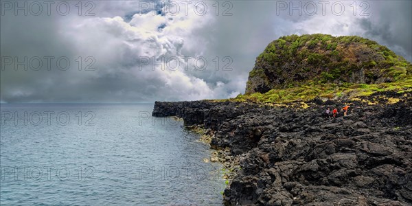 Group of people walking along a rocky coast with a cloudy sky above, lava rocks coastal path Ponta da Iiha, Calhau, west coast, Pico, Azores