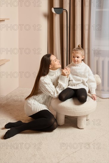 A young mother sits on the floor next to her little daughter sitting on a soft chair