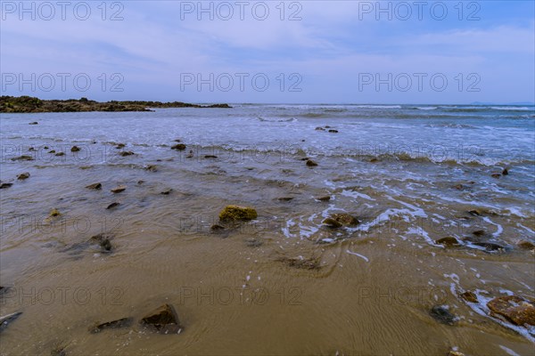 Landscape of ocean shoreline with waves washing up on rocks in the sand and a small rocky outcrop in the background