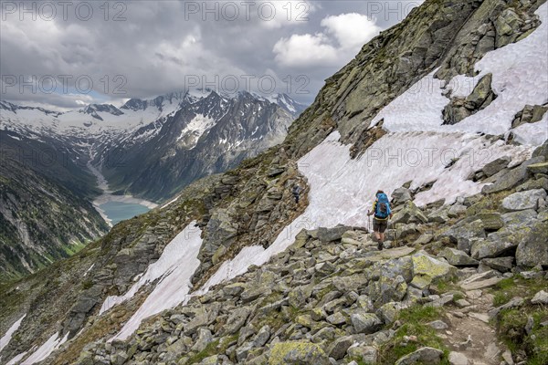 Two mountaineers on hiking trail with snow, view of Schlegeisspeicher, glaciated rocky mountain peaks Hoher Weisszint and Hochfeiler with glacier Schlegeiskees, Berliner Hoehenweg, Zillertal Alps, Tyrol, Austria, Europe