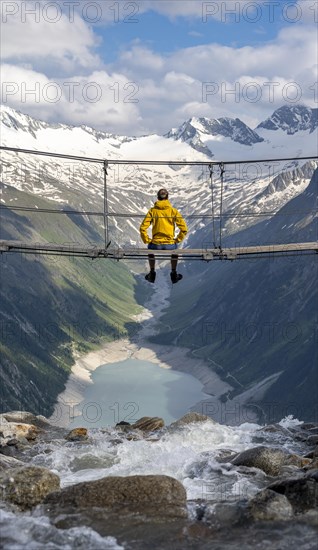 Mountaineer sitting on a suspension bridge over a mountain stream Alelebach, picturesque mountain landscape near the Olpererhuette, view of turquoise blue lake Schlegeisspeicher, glaciated rocky mountain peaks Hoher Weisszint and Hochfeiler with glacier Schlegeiskees, Berliner Hoehenweg, Zillertal Alps, Tyrol, Austria, Europe