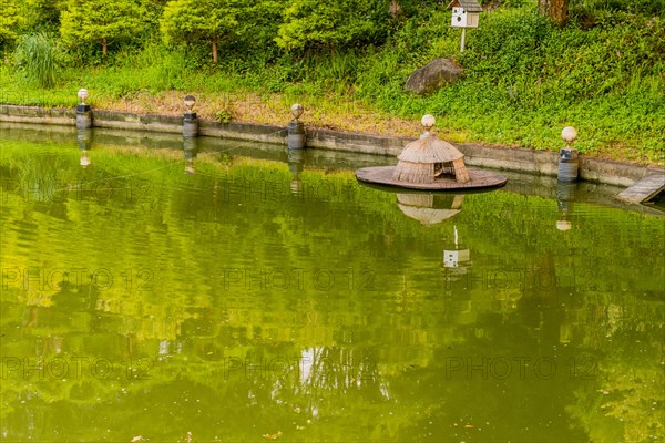 Straw duck house floating at edge of pond and birdhouse in background in public park