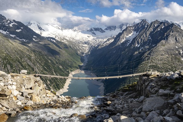 Suspension bridge over a mountain stream Alelebach, picturesque mountain landscape near the Olpererhuette, view of turquoise-blue lake Schlegeisspeicher, glaciated rocky mountain peaks Grosser Moeseler, Hoher Weisszint and Hochfeilermit glacier Schlegeiskees, Berliner Hoehenweg, Zillertal Alps, Tyrol, Austria, Europe