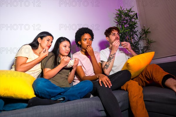 Multiracial friends watching an interesting film on tv sitting on the sofa eating popcorn