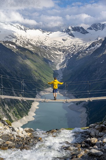 Mountaineers on a suspension bridge over a mountain stream Alelebach, picturesque mountain landscape near the Olpererhuette, view of turquoise-blue lake Schlegeisspeicher, glaciated rocky mountain peaks Hoher Weisszint and Hochfeiler with glacier Schlegeiskees, Berliner Hoehenweg, Zillertal Alps, Tyrol, Austria, Europe