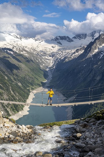 Mountaineers on a suspension bridge over a mountain stream Alelebach, picturesque mountain landscape near the Olpererhuette, view of turquoise-blue lake Schlegeisspeicher, glaciated rocky mountain peaks Grosser Moeseler, Hoher Weisszint and Hochfeilermit glacier Schlegeiskees, Berliner Hoehenweg, Zillertal Alps, Tyrol, Austria, Europe