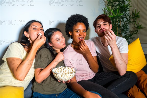 Multi-ethnic friends in a movie night at home with popcorn sitting on the sofa