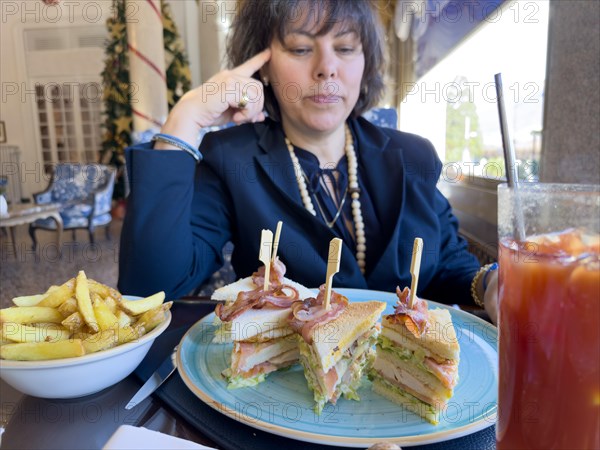 Elegant Woman Sitting Around a Table in Restaurant and Eating Club Sandwich with Bacon and French Fries and a Bloody Mary Drink in a Sunny Day in Switzerland