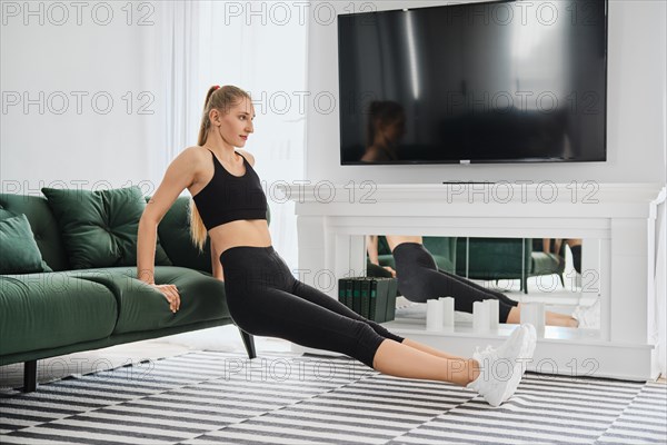 Young woman in sportswear does triceps muscle exercises at home leaning on sofa