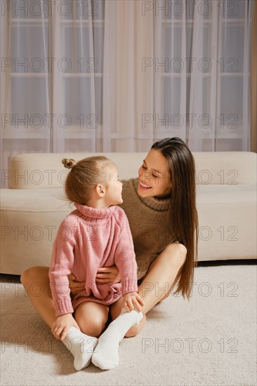 A young mother and daughter sit on the floor in a cozy living room, with a couch and curtains in the background. They wear warm knitted sweaters and socks