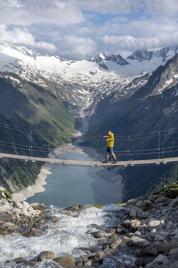 Mountaineers on a suspension bridge over a mountain stream Alelebach, picturesque mountain landscape near the Olpererhuette, view of turquoise-blue lake Schlegeisspeicher, glaciated rocky mountain peaks Grosser Moeseler, Hoher Weisszint and Hochfeiler with glacier Schlegeiskees, Berliner Hoehenweg, Zillertal Alps, Tyrol, Austria, Europe