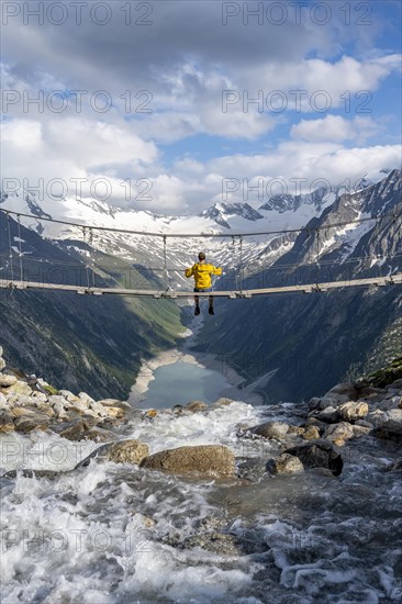 Mountaineer sitting on a suspension bridge over a mountain stream Alelebach, picturesque mountain landscape near the Olpererhuette, view of turquoise blue lake Schlegeisspeicher, glaciated rocky mountain peaks Hoher Weisszint and Hochfeiler with glacier Schlegeiskees, Berliner Hoehenweg, Zillertal Alps, Tyrol, Austria, Europe