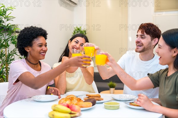 Joyful young multi-ethnic friends toasting with orange juice having breakfast at home