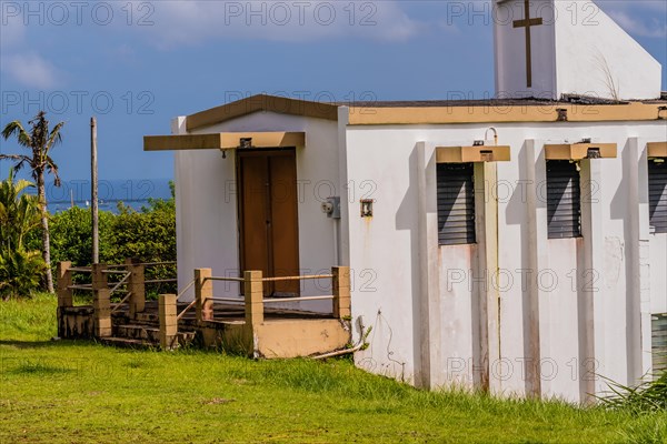 Side view of a small white country church sitting on a hillside in Guam with a view of the ocean under a cloudy sky in the background