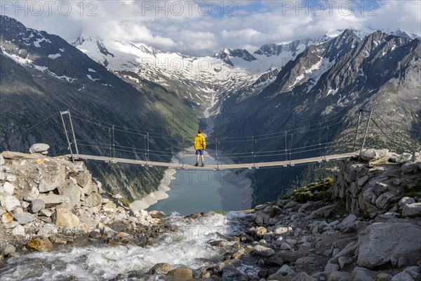 Mountaineers on a suspension bridge over a mountain stream Alelebach, picturesque mountain landscape near the Olpererhuette, view of turquoise-blue lake Schlegeisspeicher, glaciated rocky mountain peaks Hoher Weisszint and Hochfeiler with glacier Schlegeiskees, Berliner Hoehenweg, Zillertal Alps, Tyrol, Austria, Europe