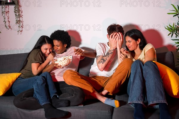 Multi-ethnic friends on a terror movie night at home sitting on the sofa eating popcorn and covering faces