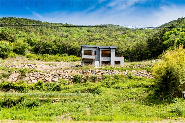 Unfinished abandoned gray concrete house in the side of a hill with stone wall in the foreground and blue sky in the background