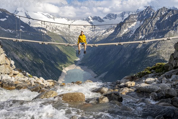 Mountaineer sitting on a suspension bridge over a mountain stream Alelebach, picturesque mountain landscape near the Olpererhuette, view of turquoise blue lake Schlegeisspeicher, glaciated rocky mountain peaks Hoher Weisszint and Hochfeiler with glacier Schlegeiskees, Berliner Hoehenweg, Zillertal Alps, Tyrol, Austria, Europe