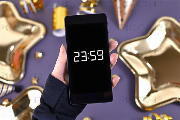Concept for New Year Silvester celebration with hand holding smart phone with timer countdown to midnight in front of party items in background