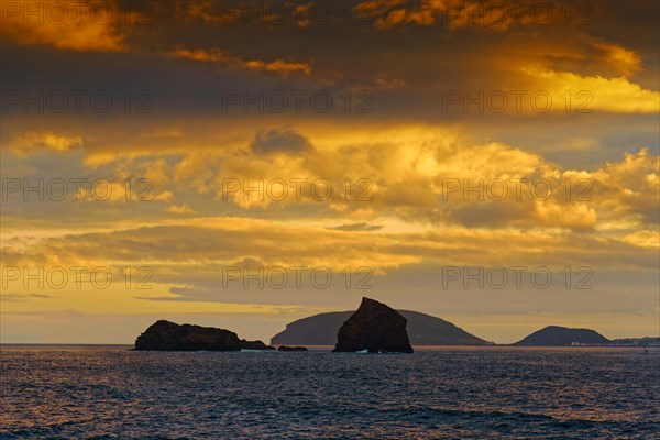 Sea view of the rocks Ilheu em Pe and Ilheu Deitado under an amber-coloured evening sky with clouds, Madalena, Pico, Azores, Portugal, Europe
