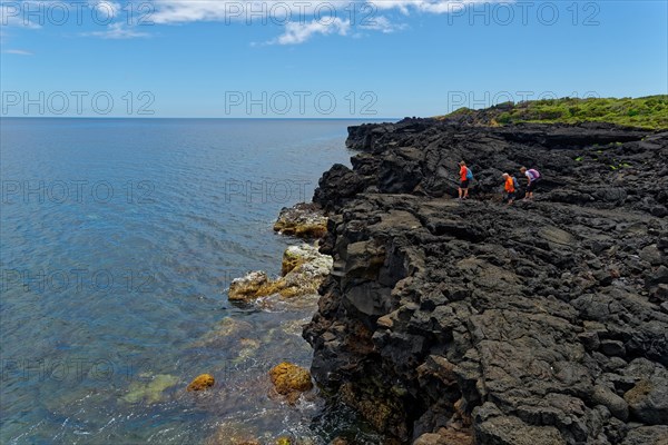 Three people hiking on rocky coastline next to the clear blue sea water, lava rocks coastal hiking trail Ponta da Iiha, Calhau, west coast, Pico, Azores