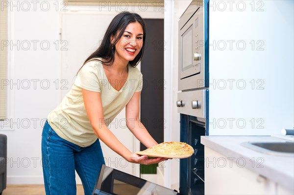 Smiling woman looking at camera while putting a pizza into a oven at home