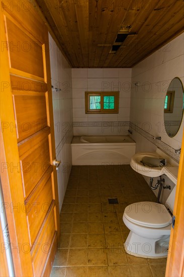 Grungy bathroom with wooden ceiling, bathtub, sink, commode and round mirror in abandoned house