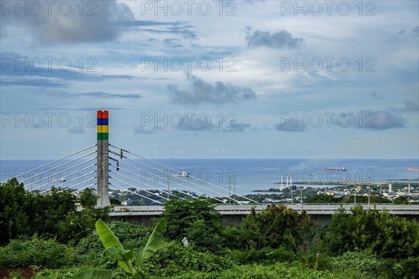 A highway with Mauritian emblem on pillar and a truck driving, surrounded by mountains and an overcast sky.A1-M1 Link Bridge At Grand River North West Valley on the island of Mauritius