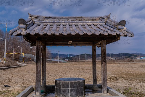 Covered water well under ceramic tiled roof full of water in a mountainside public park with plowed field in background