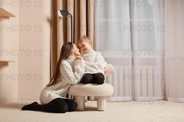 A young mother looks lovingly at her little daughter sitting on soft chair