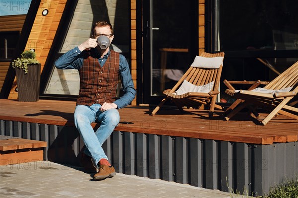 Middle-aged man sitting on terrace of wooden cabin and drinking tea in the morning