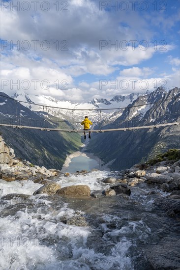 Mountaineer sitting on a suspension bridge over a mountain stream Alelebach, picturesque mountain landscape near the Olpererhuette, view of turquoise blue lake Schlegeisspeicher, glaciated rocky mountain peaks Hoher Weisszint and Hochfeiler with glacier Schlegeiskees, Berliner Hoehenweg, Zillertal Alps, Tyrol, Austria, Europe