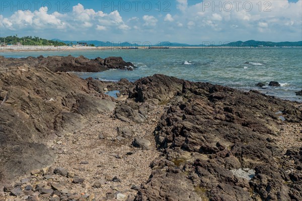 Seascape of rocking shore with beach and harbor road in background under cloudy blue sky