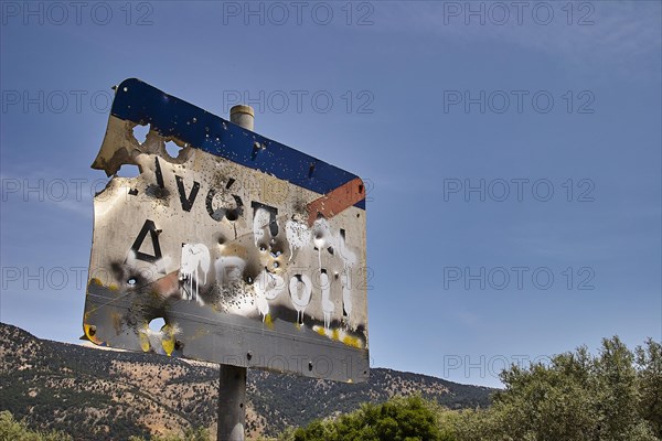 Weathered road sign with bullet holes against a blue sky and trees in the background, Anopolis, Sfakia, West Crete, Crete, Greece, Europe