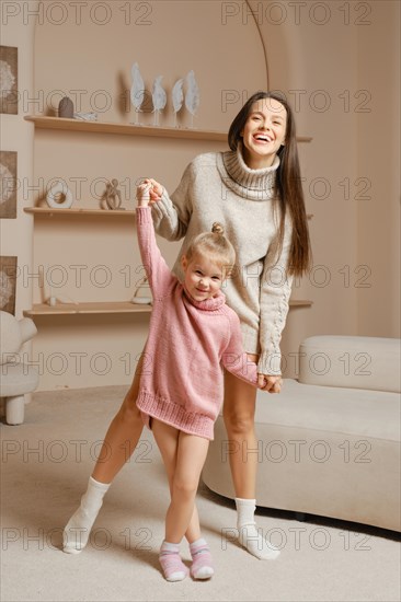 A mother and her young daughter laughing while playing together at home. They both wear knitted sweaters and warm socks