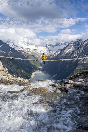 Mountaineer sitting on a suspension bridge over a mountain stream Alelebach, picturesque mountain landscape near the Olpererhuette, view of turquoise blue lake Schlegeisspeicher, glaciated rocky mountain peaks Hoher Weisszint and Hochfeiler with glacier Schlegeiskees, Berliner Hoehenweg, Zillertal Alps, Tyrol, Austria, Europe