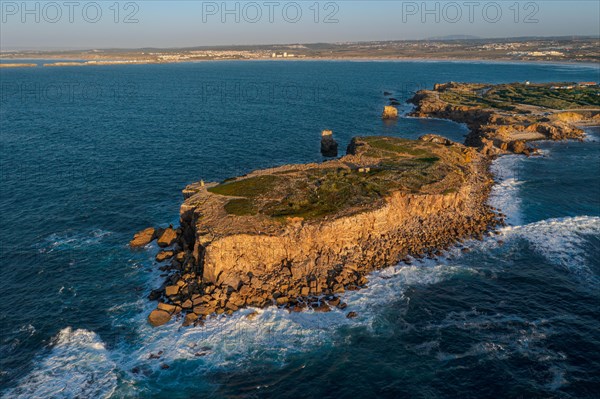Aerial image of high rocky headland and peninsula. Drone shot of sea blue waves beat and splash in summer sunset haze, little foliage and rocky cliffs, establishing or static shot, Peniche, Portugal, Europe