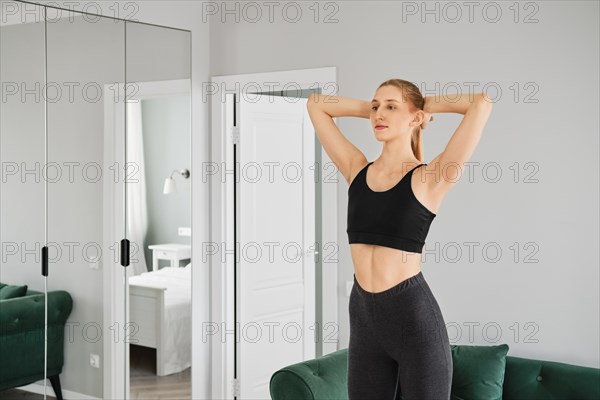 Young sporty woman warming up in her bright apartment doing turns and bends with her arms behind head