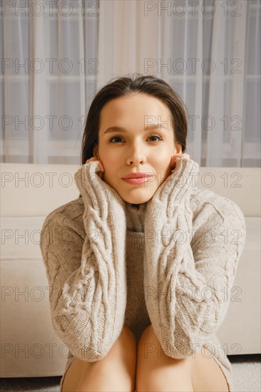 A contemplative woman, wrapped in a cozy knitted sweater, sits next to bed. Her hands wrap around her face, and the room is bathed in soft light