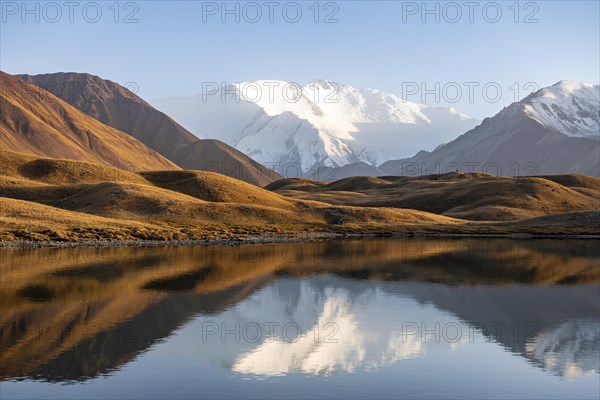 White glaciated and snowy mountain peak Pik Lenin at sunset, mountains reflected in a lake between golden hills, Trans Alay Mountains, Pamir Mountains, Osh Province, Kyrgyzstan, Asia