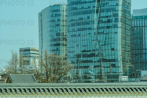 Tall buildings with glass exteriors with a wall and the roof of a small building made in traditional Korean style archetecture in the foreground