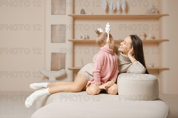 A mother and daughter sit together in a cozy living room with a beige walls. The daughter is wearing a pink sweater and a hair hoop