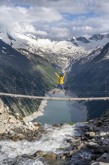 Mountaineer jumping on a suspension bridge over a mountain stream Alelebach, picturesque mountain landscape near the Olpererhuette, view of turquoise blue lake Schlegeisspeicher, glaciated rocky mountain peaks Hoher Weisszint and Hochfeiler with glacier Schlegeiskees, Berliner Hoehenweg, Zillertal Alps, Tyrol, Austria, Europe