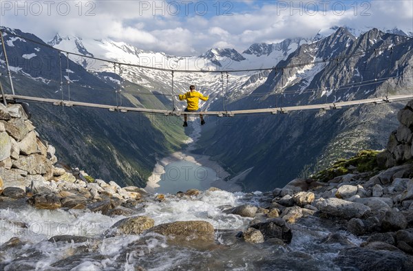 Mountaineer sitting on a suspension bridge over a mountain stream Alelebach, picturesque mountain landscape near the Olpererhuette, view of turquoise blue lake Schlegeisspeicher, glaciated rocky mountain peaks Hoher Weisszint and Hochfeiler with glacier Schlegeiskees, Berliner Hoehenweg, Zillertal Alps, Tyrol, Austria, Europe